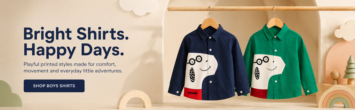 Two long-sleeve button-up shirts for boys hang on wooden hangers against a soft cream background. The left shirt is navy blue with a white cartoon face and red pants graphic, and the right shirt is green with a white cartoon face and red pants graphic.